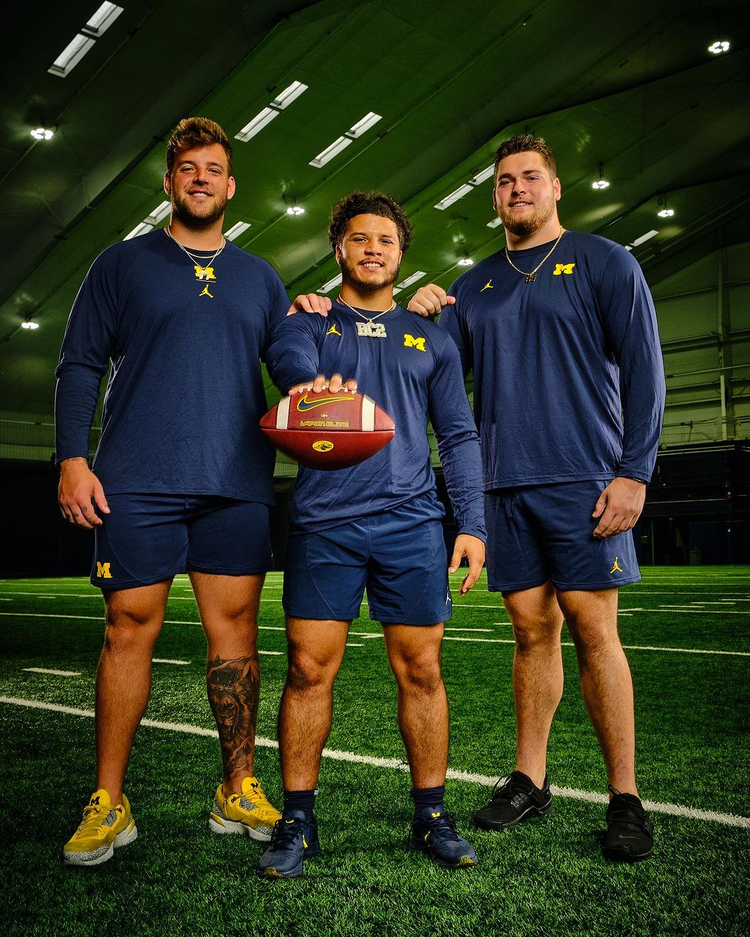 Three football players in Michigan Wolverines uniforms pose on a turf field inside an indoor training facility. They hold a football.