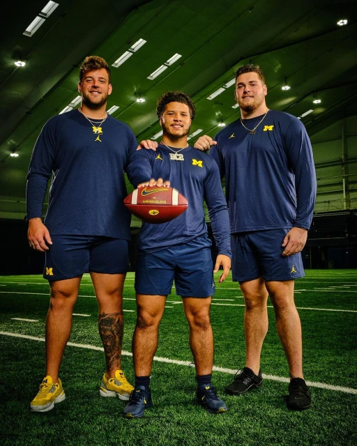 Three football players in Michigan Wolverines uniforms pose on a turf field inside an indoor training facility. They hold a football.