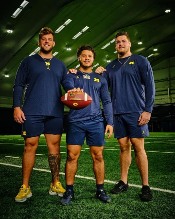 Three football players in Michigan Wolverines uniforms pose on a turf field inside an indoor training facility. They hold a football.