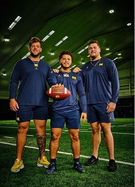 Three football players wearing uniforms and holding a football pose indoors on a green surface with a green background and bright lights.