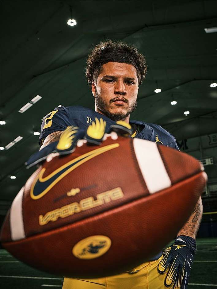 Male football player in uniform poses indoors holding football with logo in eye-level shot with diffused natural lighting.