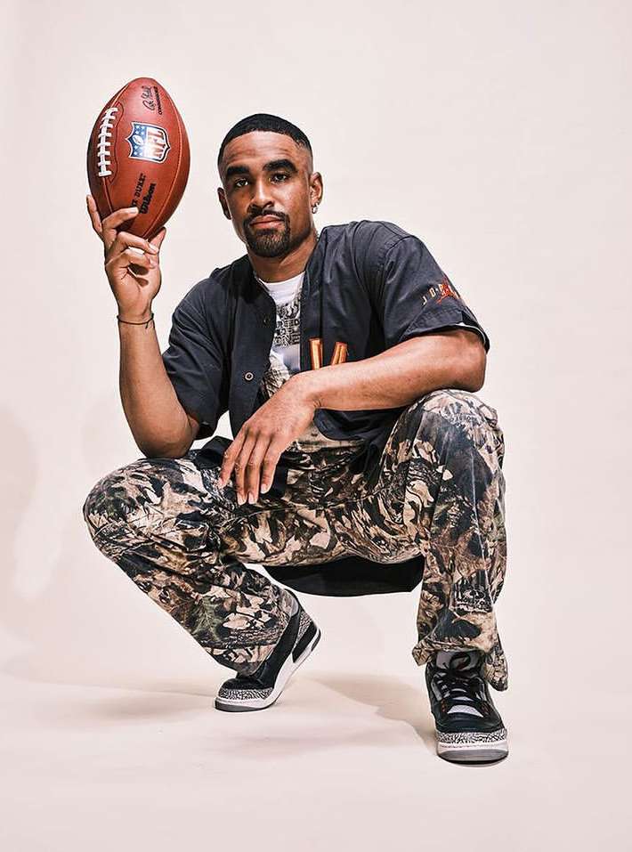 Adult male athlete posing in a studio setting while holding a football with camo pants, baseball shirt, and sneakers.