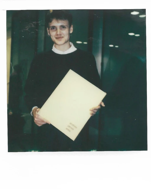 A young adult man stands indoors holding a large, light yellow portfolio with visible text against a dark-green glass-paneled wall background.