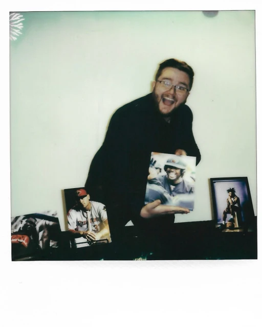 Enthusiastic man in glasses holds up a photo; additional framed photos sit on a surface in the background against a white wall.