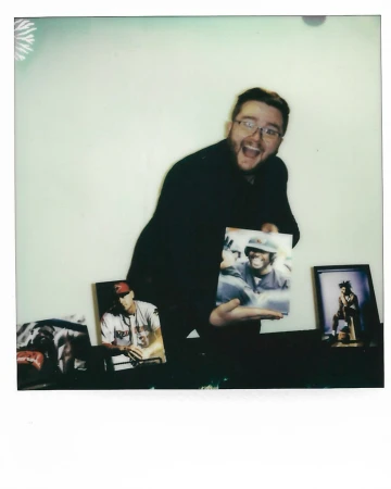 Enthusiastic man in glasses holds up a photo; additional framed photos sit on a surface in the background against a white wall.