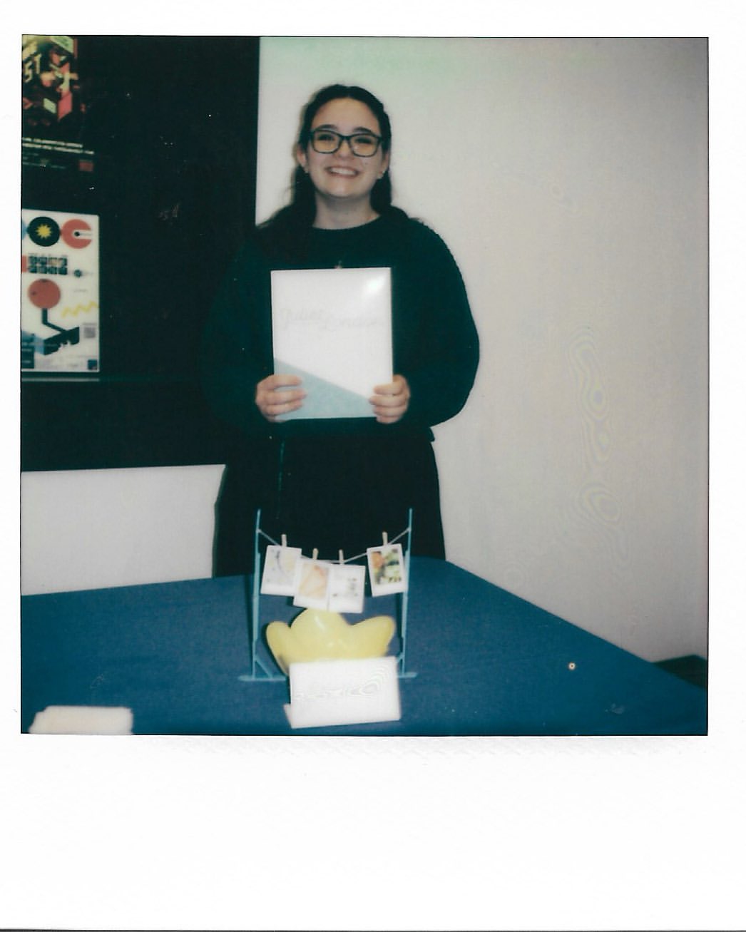 A young adult woman with glasses stands behind a blue table and presents a white book or album cover in an indoor setting.