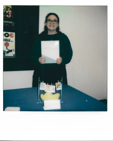 A young adult woman with glasses stands behind a blue table and presents a white book or album cover in an indoor setting.