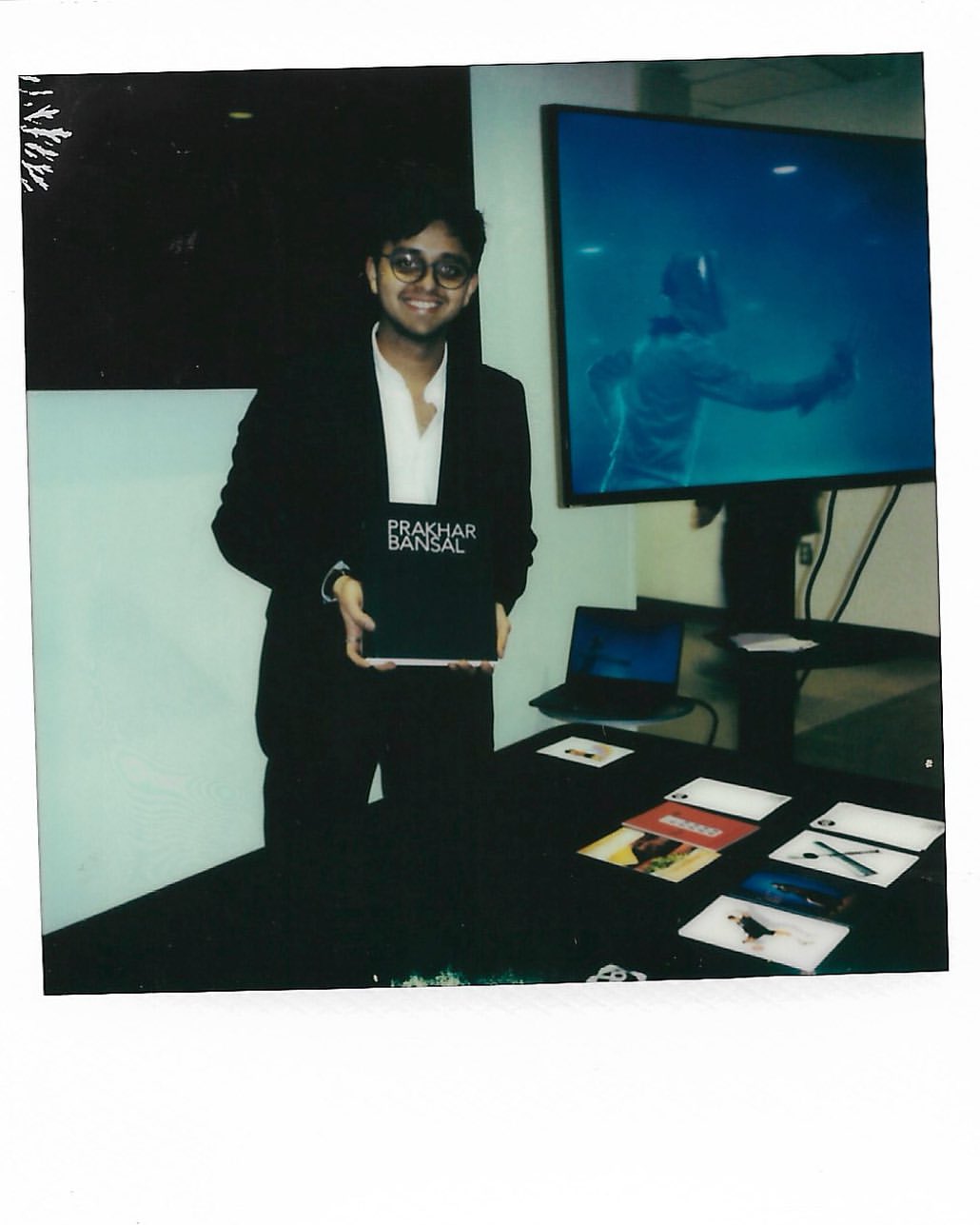 Adult male in a suit poses indoors holding a book in front of a table with cards and a laptop, with a video monitor in the background.