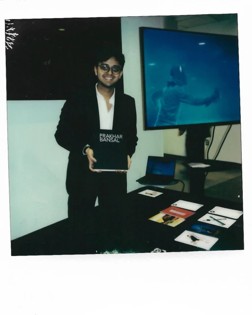 Adult male in a suit poses indoors holding a book in front of a table with cards and a laptop, with a video monitor in the background.