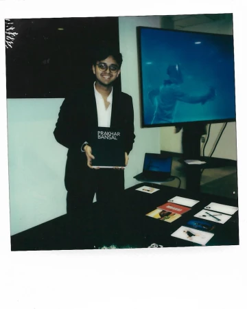 Adult male in a suit poses indoors holding a book in front of a table with cards and a laptop, with a video monitor in the background.