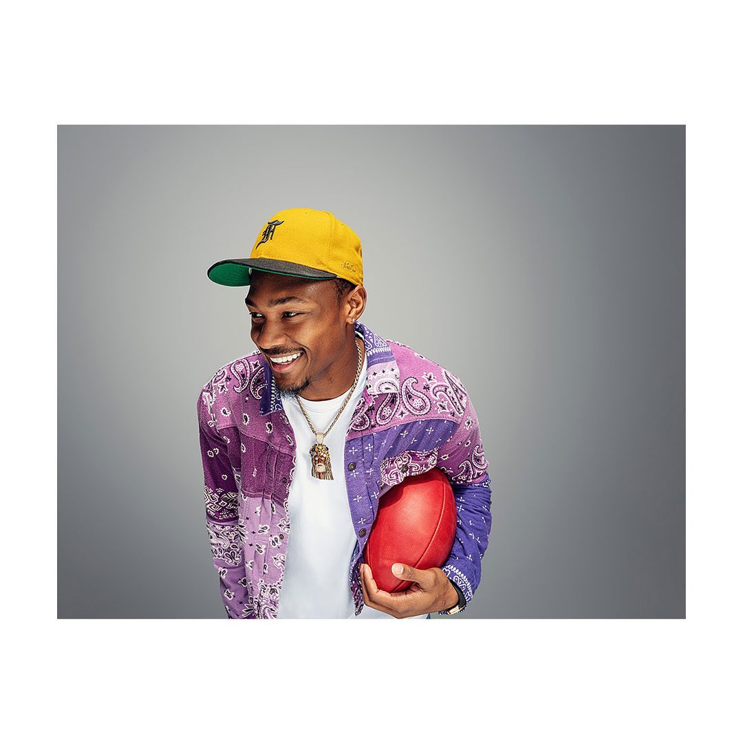 Smiling adult man with football, wearing a paisley jacket and baseball cap, in a studio shot against a grey background.