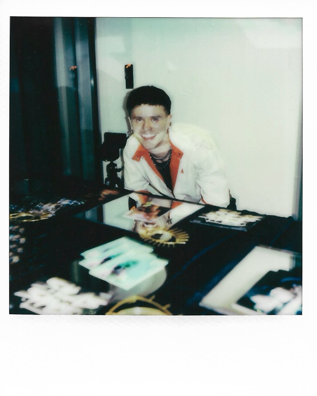 Person smiling in Polaroid style indoor shot, showcasing fashion jacket, jewelry and cards on table in soft diffused lighting.