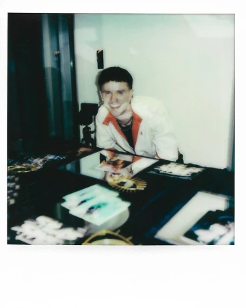 Person smiling in Polaroid style indoor shot, showcasing fashion jacket, jewelry and cards on table in soft diffused lighting.