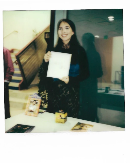 Smiling woman holds white paper above table with printed photos and a small yellow box. The scene is indoors and the woman is center frame.