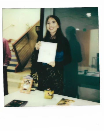 Smiling woman holds white paper above table with printed photos and a small yellow box. The scene is indoors and the woman is center frame.