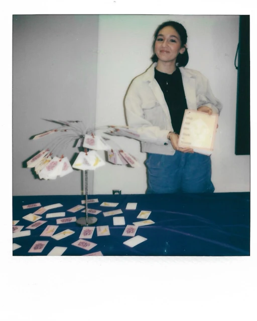 Young woman stands behind table covered with cards, holding a covered book. A card display is visible. Indoors. Studio lighting.