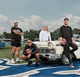 Four men posing outdoors near a vintage white convertible car on an athletic field under a partly cloudy sky.