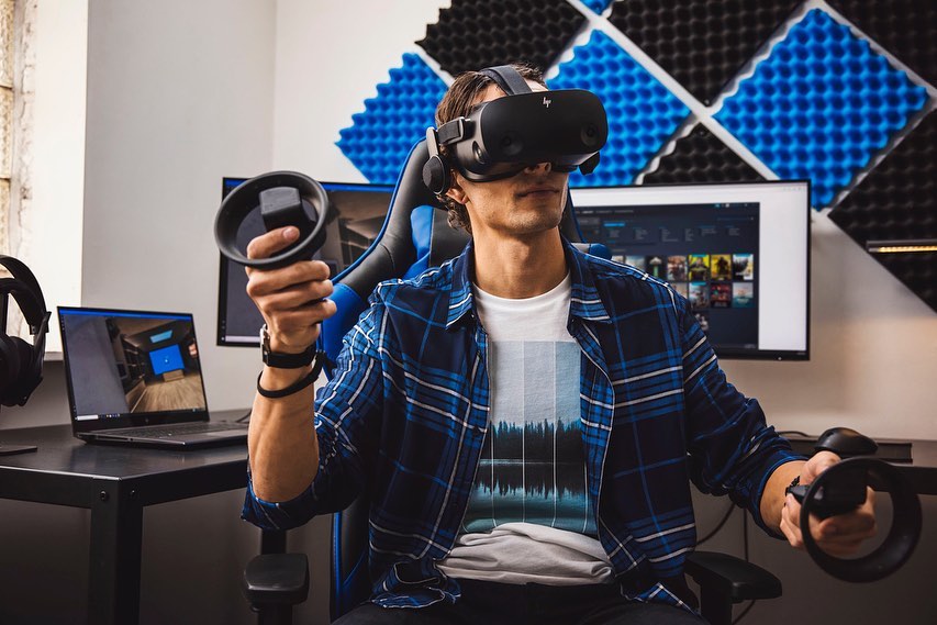 Man wearing a VR headset and holding controllers while gaming on a gaming chair in an indoor environment with computer and peripherals.