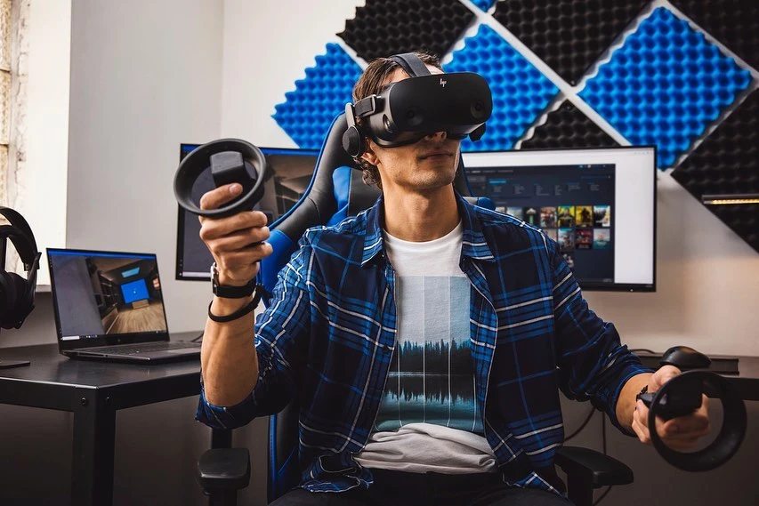Man wearing a VR headset and holding controllers while gaming on a gaming chair in an indoor environment with computer and peripherals.