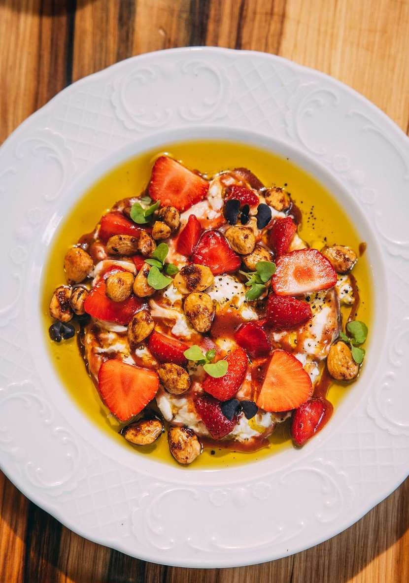 Overhead shot of a white plate with fresh strawberries, nuts, microgreens, and a drizzle of syrup on a wooden table.