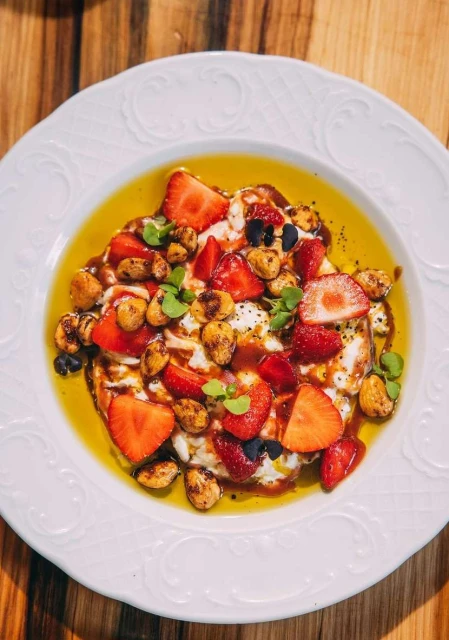 Overhead shot of a white plate with fresh strawberries, nuts, microgreens, and a drizzle of syrup on a wooden table.
