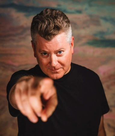 Man with grey hair wearing a black shirt points directly at the camera in a photo studio with a textured background.