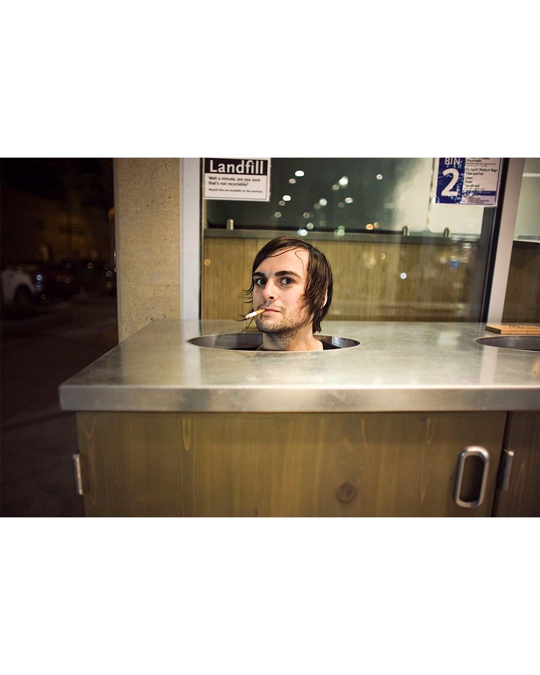 Adult man with a cigarette stands in a metal bin outdoors at night, with a partially visible sign in the background.