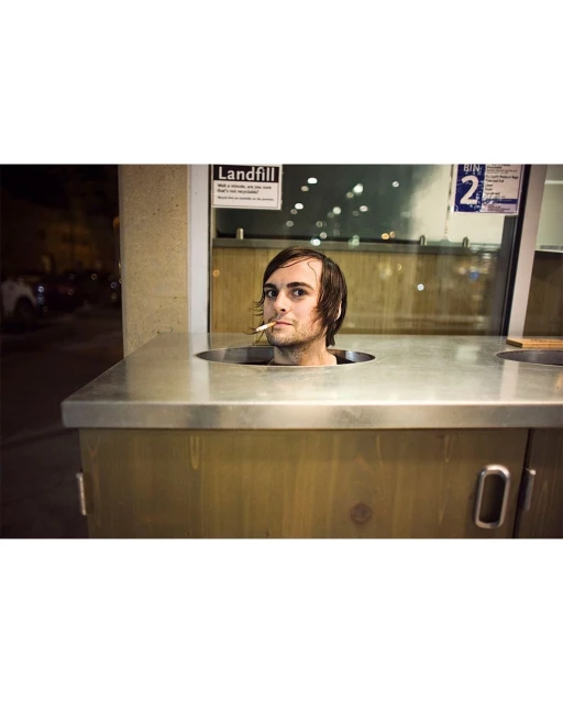 Adult man with a cigarette stands in a metal bin outdoors at night, with a partially visible sign in the background.