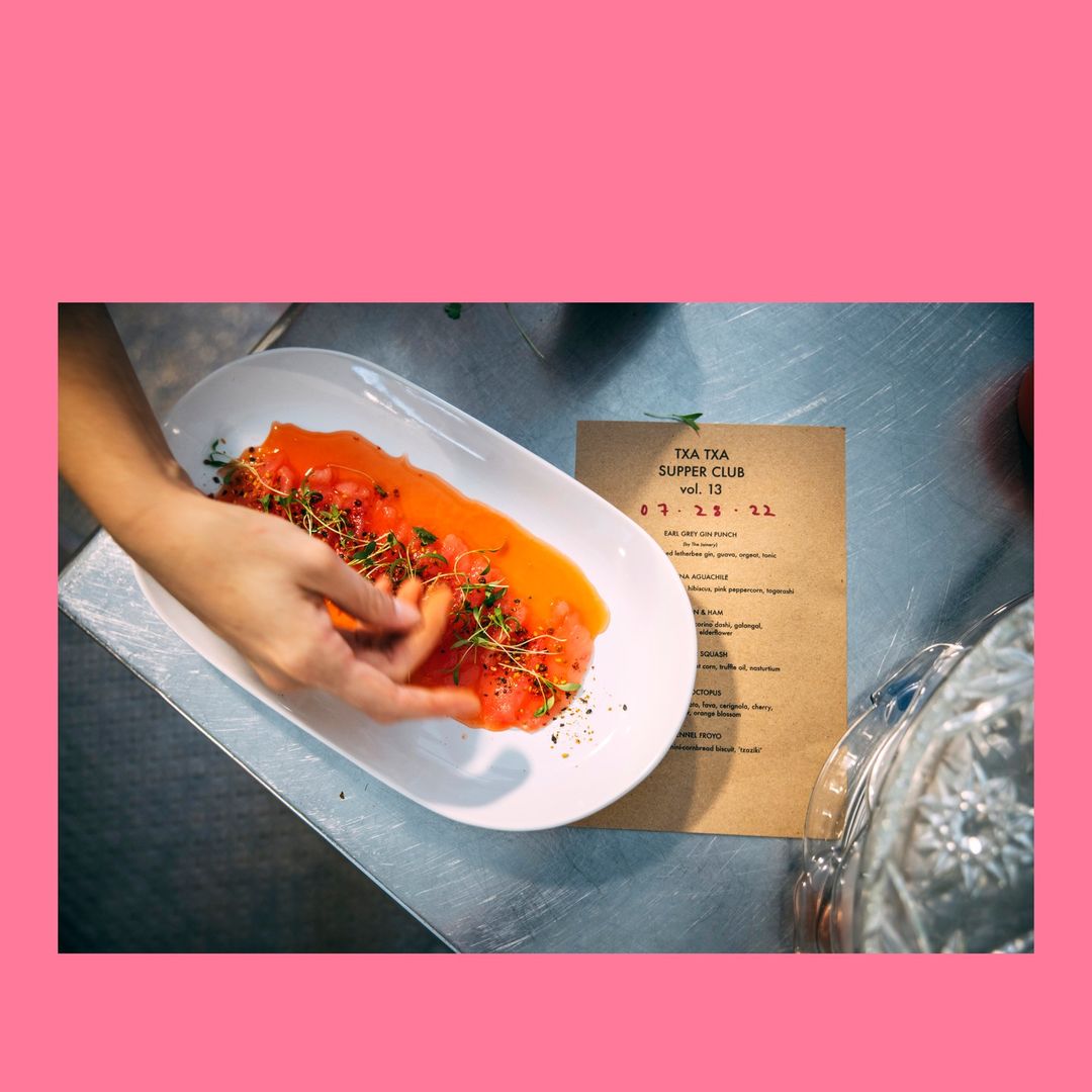 Overhead shot of hands arranging orange appetizer on a white plate, next to a menu on a stainless steel surface in a kitchen.