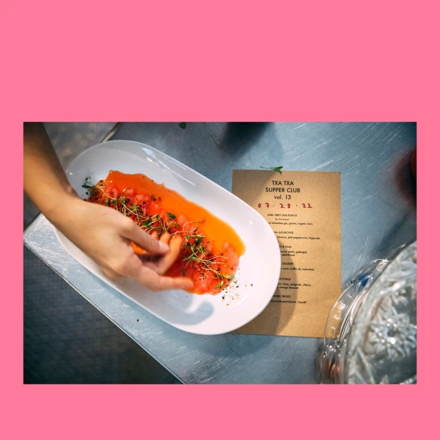 Overhead shot of hands arranging orange appetizer on a white plate, next to a menu on a stainless steel surface in a kitchen.