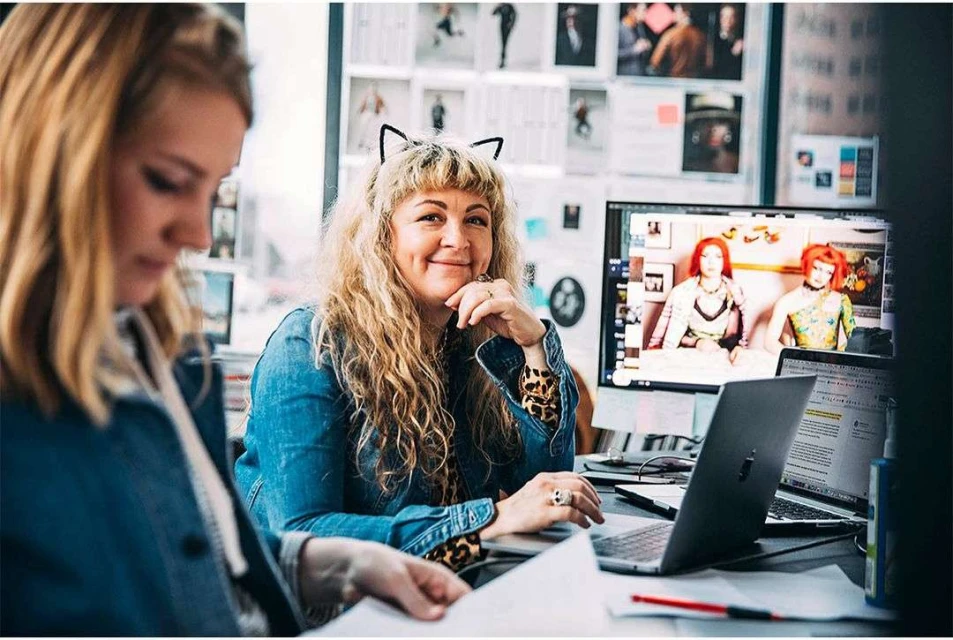 Two women work at laptops in a brightly lit office. One woman smiles at the camera wearing black cat ears and a denim jacket.