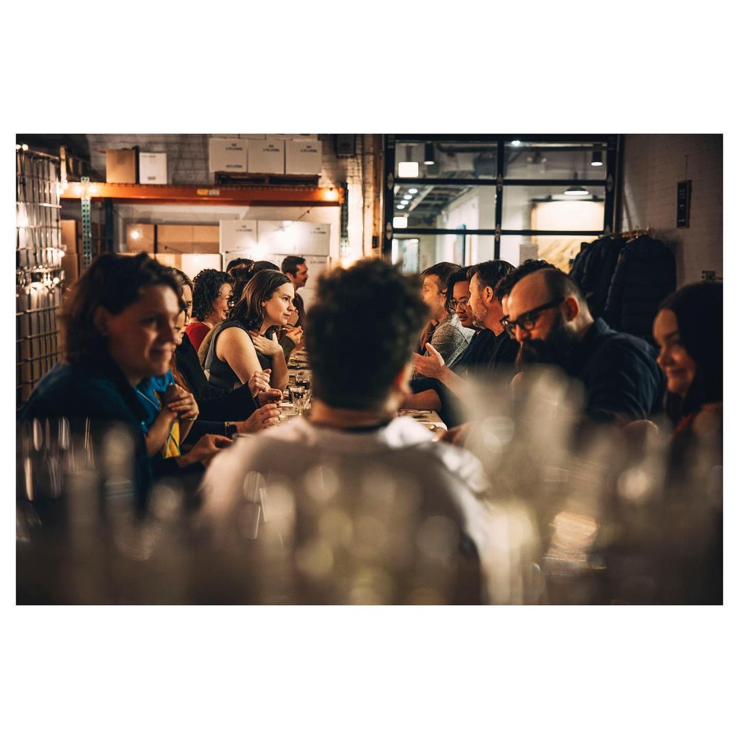 Group of people sitting at a long table, dining in a warehouse or large indoor space, glasses and plates visible, indoor lighting.