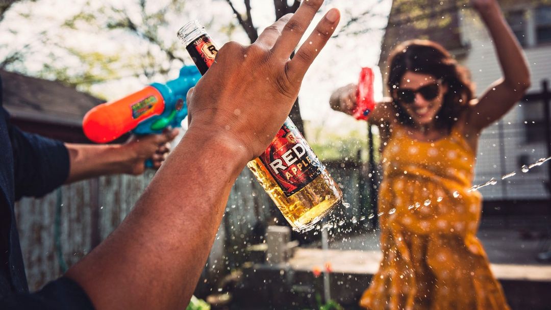 People having fun outdoors with a water fight and a Red Apple alcoholic beverage in clear glass bottle with a silver cap.