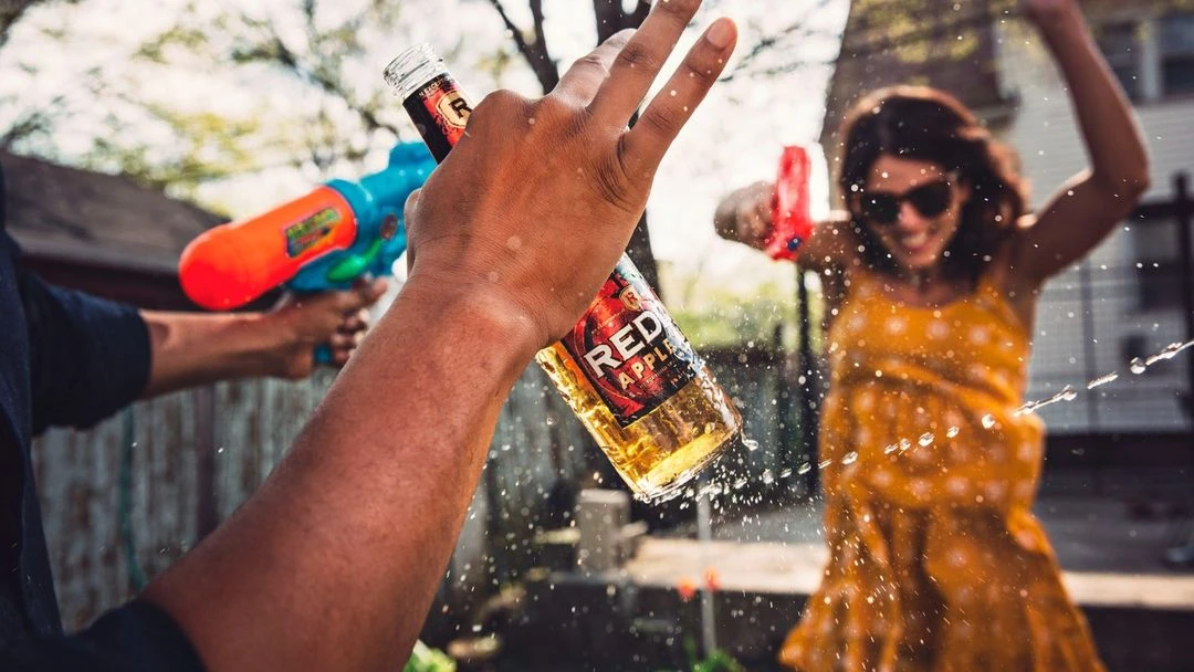 People having fun outdoors with a water fight and a Red Apple alcoholic beverage in clear glass bottle with a silver cap.