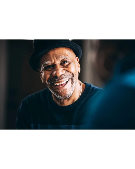 Close up of a smiling older adult male wearing a black hat and a dark navy sweater indoors, candid shot with soft, diffused lighting.