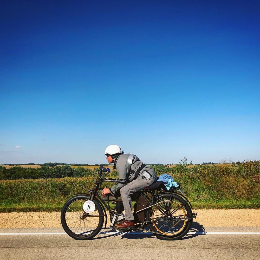 An adult male in grey clothing rides a vintage motorcycle along a rural road under a blue sky, with a field in the background.