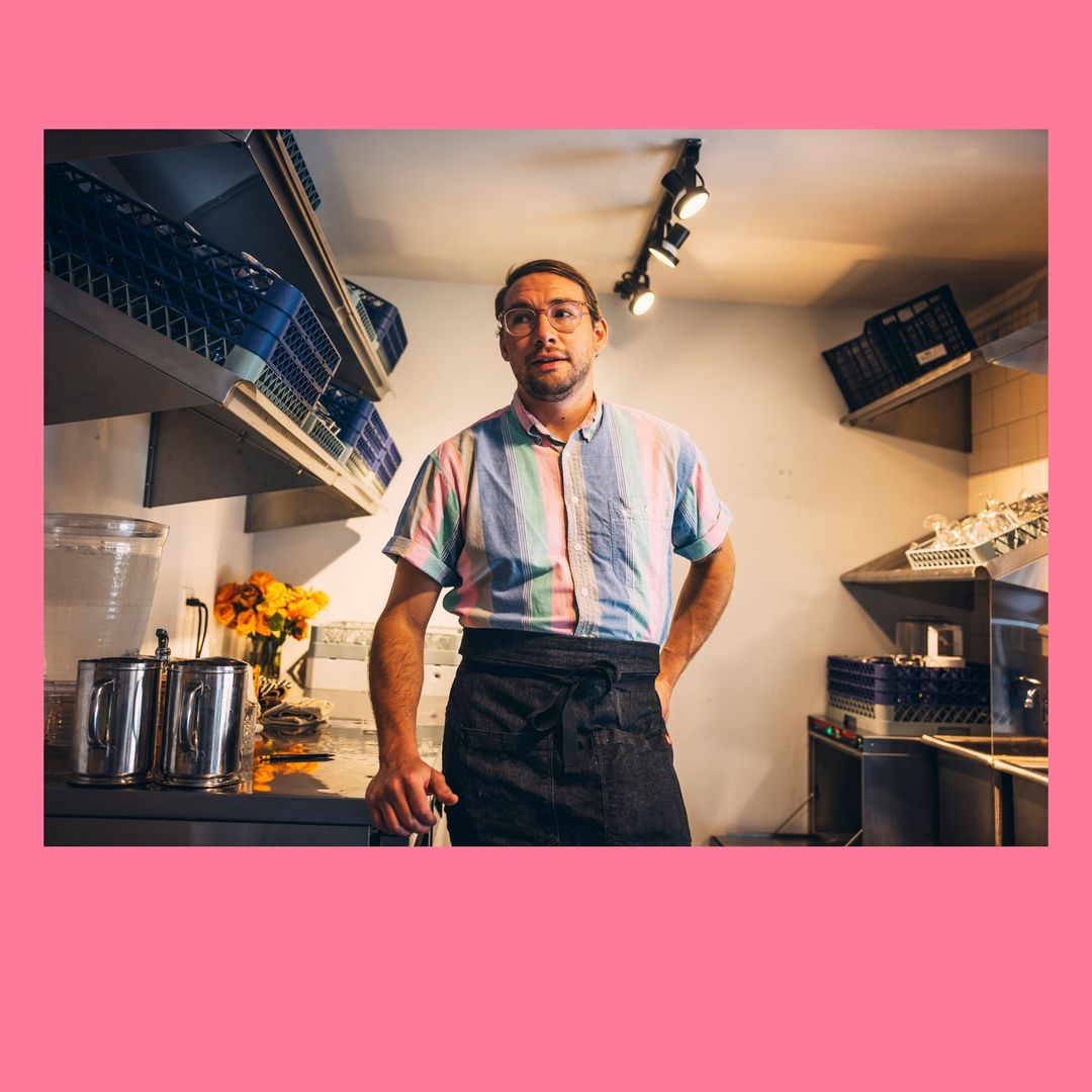 Adult male wearing a striped shirt and apron stands in a commercial kitchen with metal shelves and washing crates.