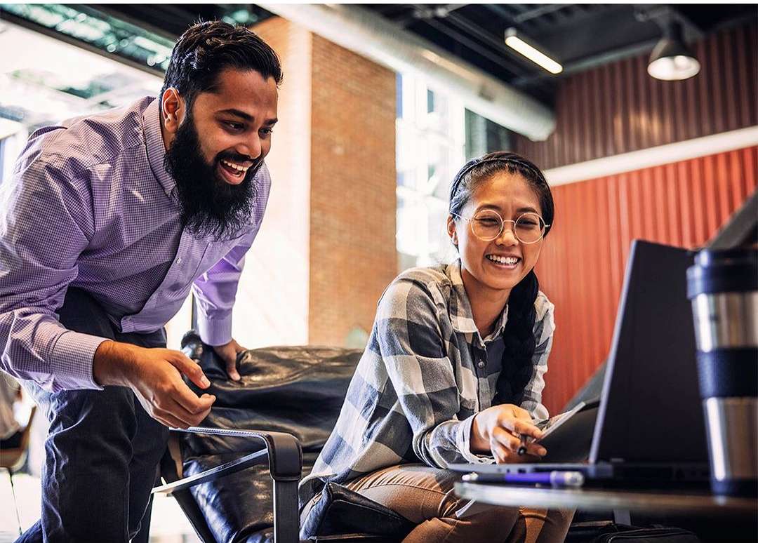 Two smiling young adults work together at a laptop in a bright, modern office space. A woman sits while a bearded man leans over.