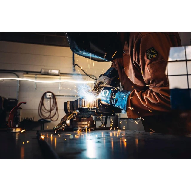 Person welding metal in a workshop with sparks flying, wearing protective gear, gloves, and a welding mask in an industrial setting.