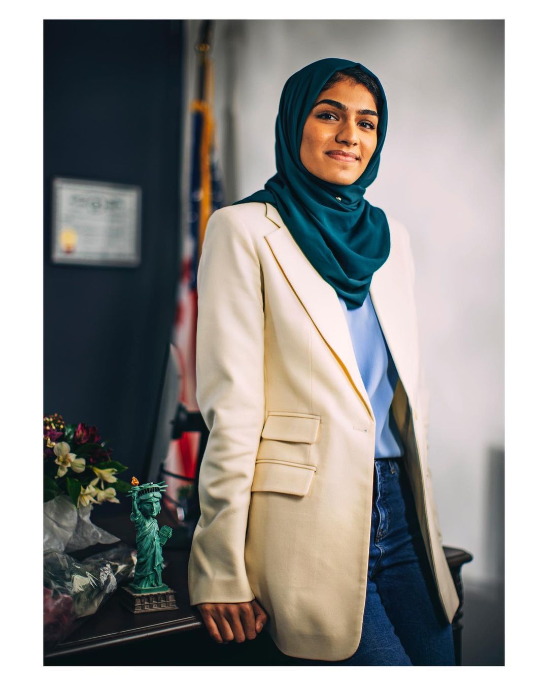 Portrait of a young adult woman wearing a teal headscarf and off-white blazer. Indoor shot in front of American flag with flowers.