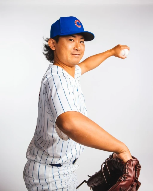 Baseball player in Chicago Cubs uniform throwing a ball, wearing a blue cap and brown baseball glove against a plain white backdrop.