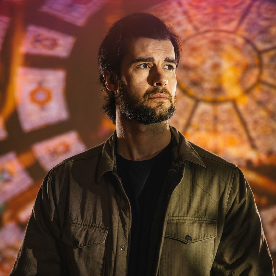 Portrait of a contemplative adult male with a beard, wearing a jacket, standing indoors with an ornate stained-glass window behind him.
