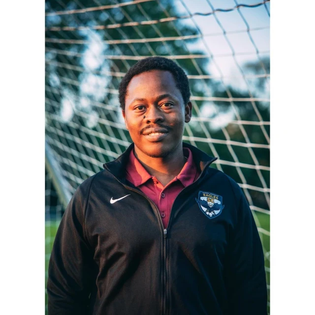 Smiling man in a soccer team jacket stands in front of a soccer net, creating a portrait with a sports theme.