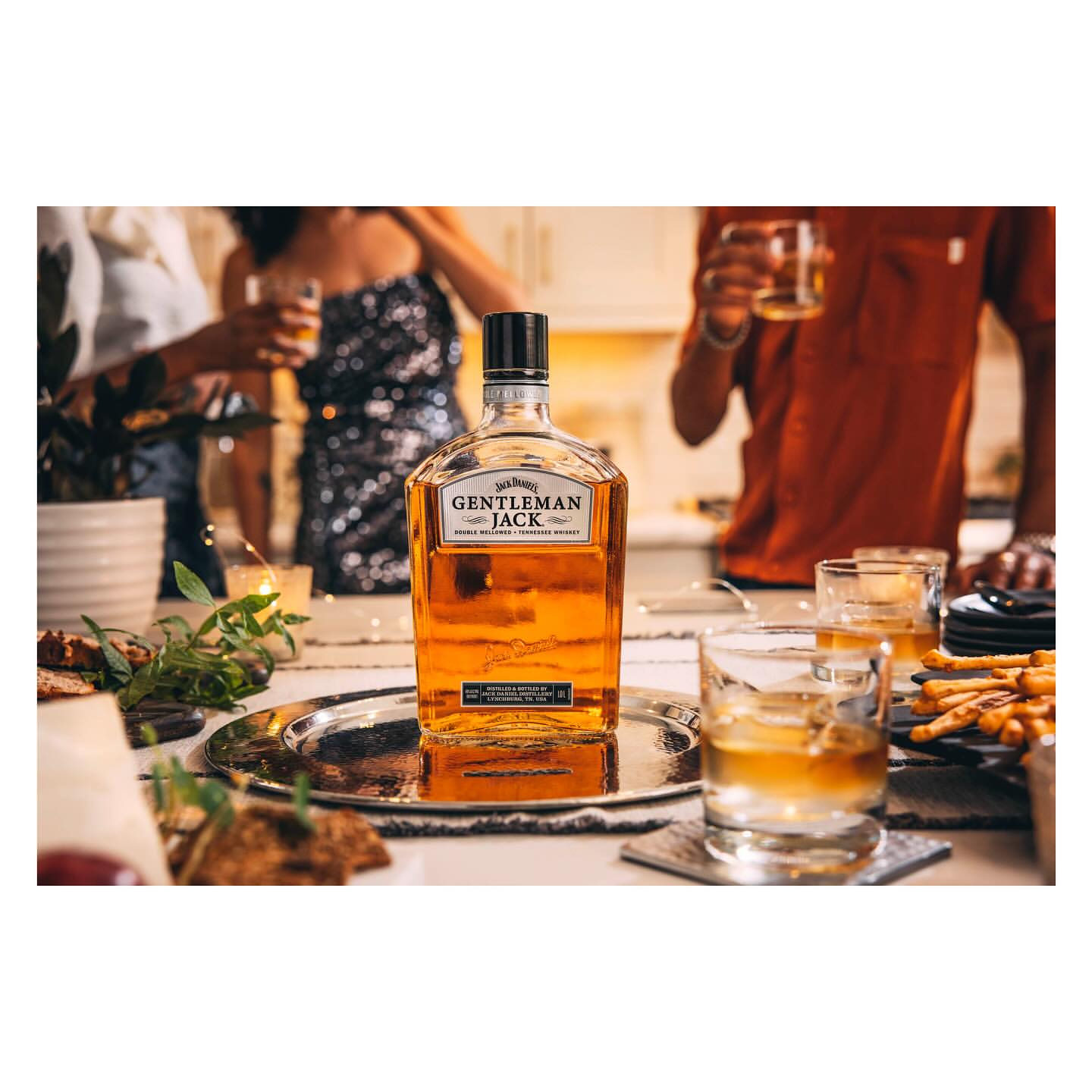 Bottle of Gentleman Jack whiskey on a table with glasses and food, with people in the background at an indoor social gathering.