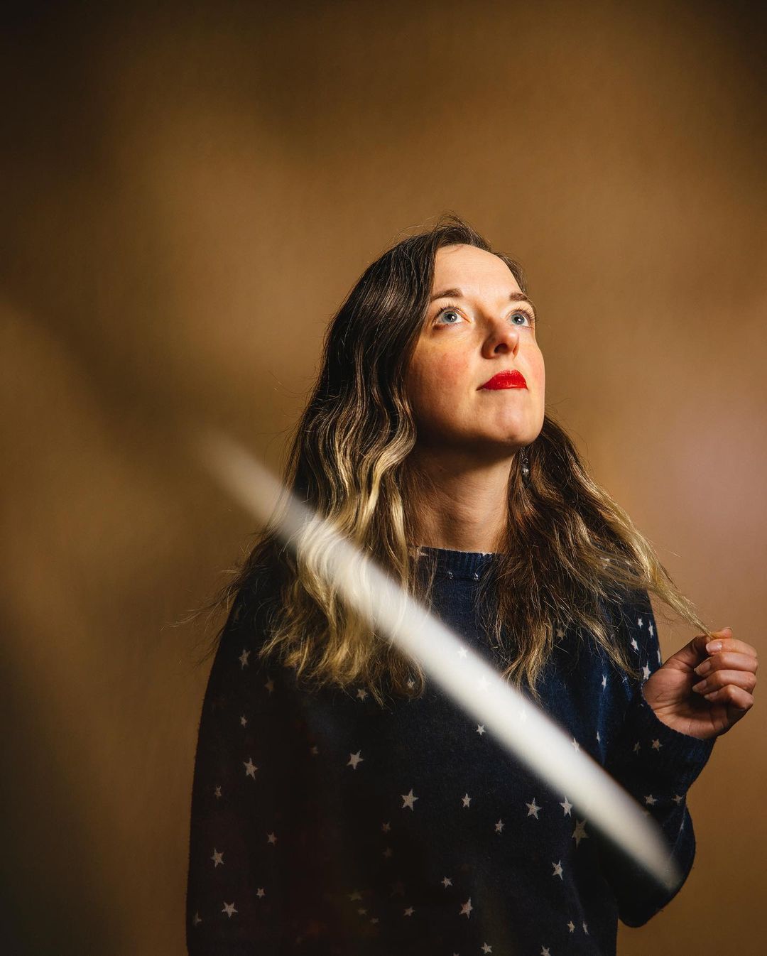 Portrait of a young woman with long, wavy hair and red lipstick looking up against a brown background with a light streak across the frame.