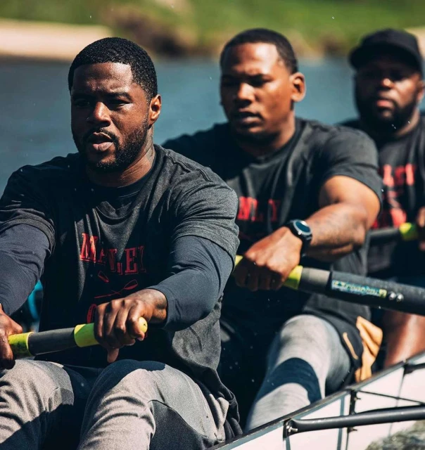 Three adult male athletes rowing a boat on a body of water under natural light, wearing matching grey t-shirts in a team activity.