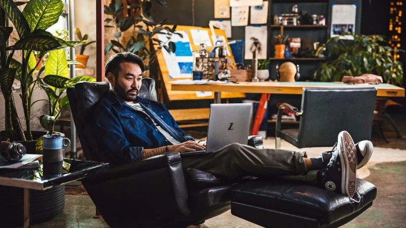 Man works on a silver laptop in a black leather chair with plants, art, and natural light in the background, creating a casual workspace.