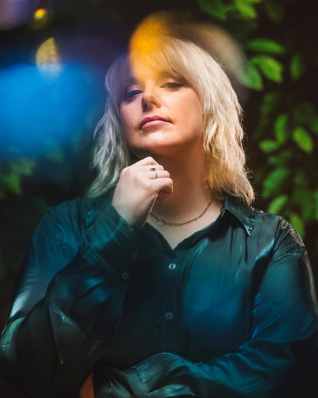 Close-up portrait of a blonde woman with her hand under her chin, wearing a green shirt and a gold necklace in front of a leafy background.