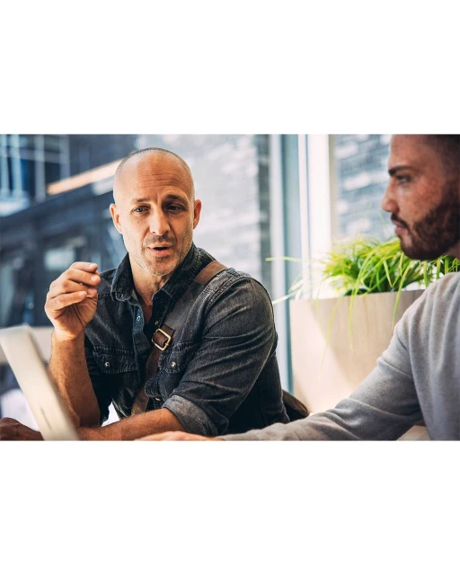 Two business men talking at a meeting table in front of a window with laptops and plants. The bald man wears a denim shirt.