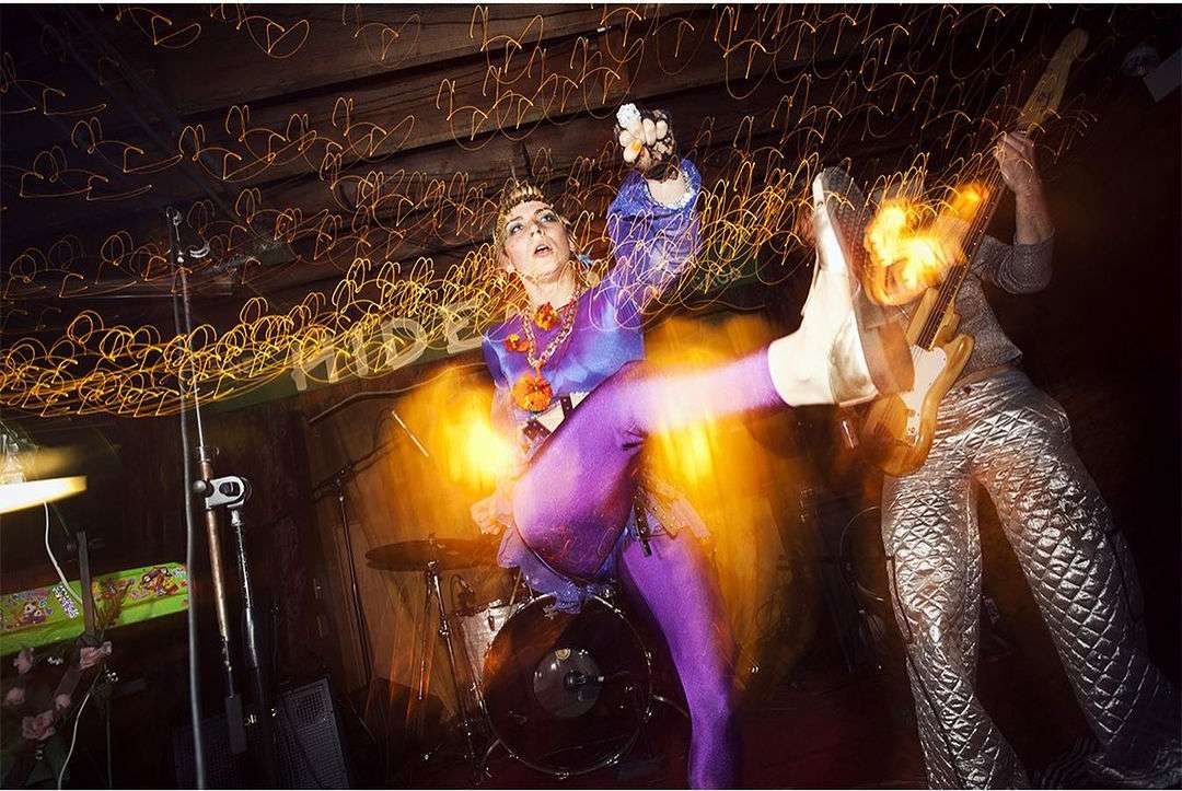 Woman in costume performs with band in a concert venue with vibrant lighting, wearing blue and purple outfit, kicking her leg in the air.